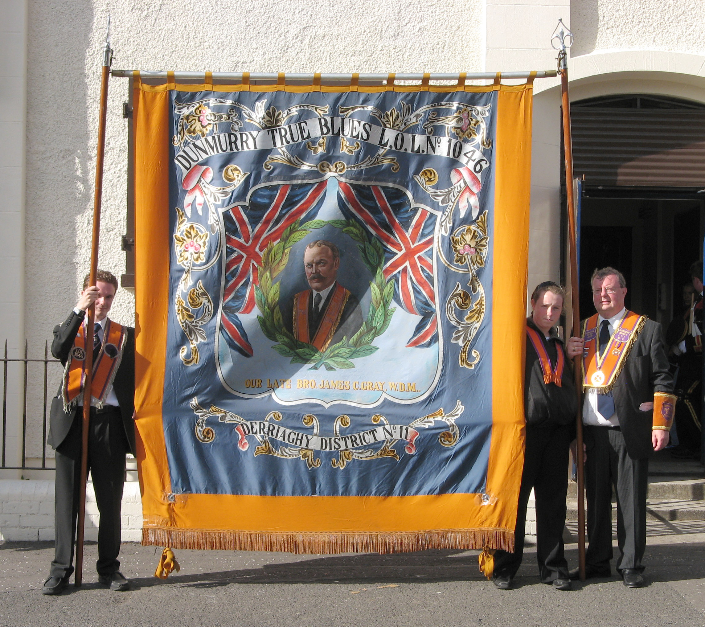Bro James Connor Gray (Worshipful District Master of Derriaghy LOL No 11) pictured with his daughter Hannah (right) and son Matthew with the banner showing a painting of his grandfather, the late Bro James Connor Gray PDM, who was present when the foundation stones of both Dunmurry Orange Hall and Saint Colmanï¿½s Parish Church in 1908 were laid.