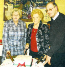 Founder members and joint chairpersons of Ballymacash Monday Club, Mrs Maud Fair and Mrs Lily Armstrong, cut the Monday Club anniversary cake under the watchful eye of Canon George Irwin.