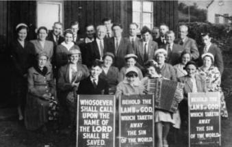 Pictured at the Assemblies of God Mission Hall in 1959 is L to R: (seated - front row) David Graham, Miss Cherry, Miss Gwen Cherry (with accordion) and Annie Kerr. (seated - second row) Mrs Bailie, Mrs Miskimmons, Mrs Lowry, Mrs Graham, Elizabeth Mulligan, Mrs Cherry and Mrs Murray. (standing) Mary McIlrath, Violet Lowry, David Graham, Leila Mulligan, Tom Scott, Sandy Miskimmons, Bob McIlrath, Will Malcomson, William Mulligan, Tommy Rowan, Aubrey Malcomson, David Murray, Mr Rogers, John Boston, Tommy McCullough and Eddie Hughes. (Thompson’s garage can be seen in the right hand side of the picture).