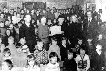 Mrs Sarah Crothers presenting a ten year savings certificate to Mrs E. McAllister, secretary of Ballymacrickett Primary School saving group in February, 1971. Looking on are the pupils with Mr T. Harvey, Miss B. Devlin, vice-principal and Mr. Jim McCann, principal.